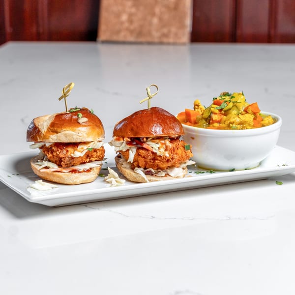 Two crispy chicken sliders on pretzel buns with coleslaw and a side of curry vegetables.