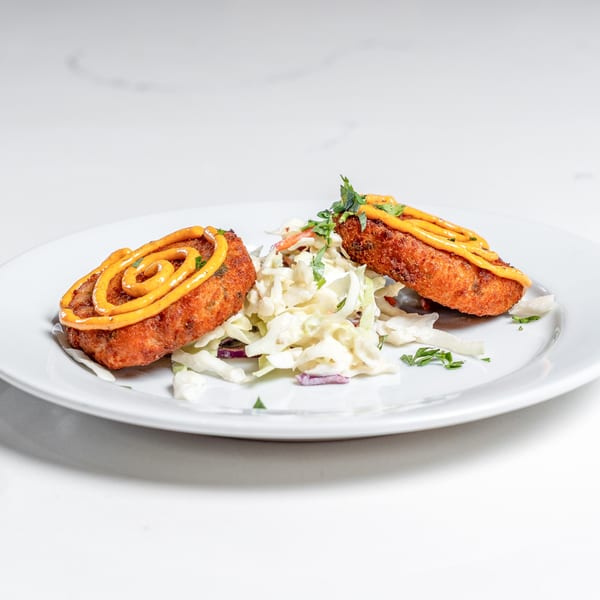 Two golden-brown crab cakes with a swirl of orange sauce, served with coleslaw on a white plate.